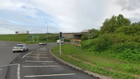 Part of the Brampton Interchange near the busy A14 and A1. There are several vehicles on the road and one on a bridge. There are traffic lights on the road and signs to exits on the road. The large roundabout has many shrubs as well as grass