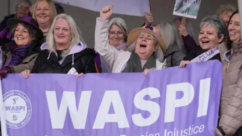 A group of older women wear purple and smile at a demonstration. They are holding signs and a purple banner that reads 'WASPI' 