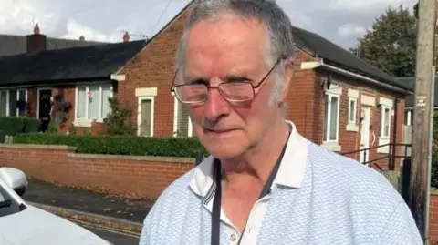 LDRS David Kelsall frowns while peering over his glasses, stood outside on a street in front of a house next to his silver electric vehicle. 