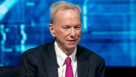 Eric Schmidt sits with his hands crossed and looks ahead. He is wearing a black suit, a white shirt, and a hot pink polka-dot tie. In the background is a blue screen that features different shades of blue.