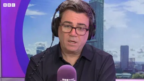 Andy Burnham sits in the Radio Manchester studio with a purple microphone in front of him, wearing glasses and over the head headphones.