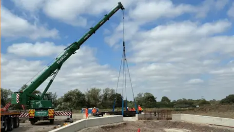 A crane being used to carry a bridge. Workers in high-vis jackets surround it.