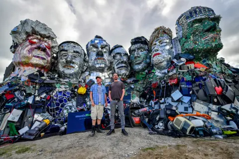 Ben Birchall / PA Media Artists Alex Wreckage, left, and Joe Rush, right, stand in front of their artwork Mount Trashmore, on the beach near Gwithian