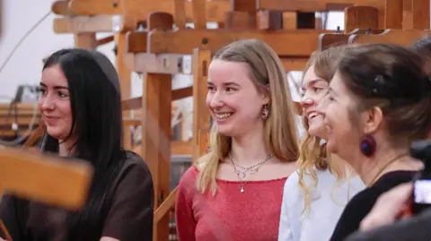 Heriot-Watt University A group of three young women stands smiling - one has dark hair, they other two have lighter blonde hair. Another older woman is closest to the camera with dark hair.