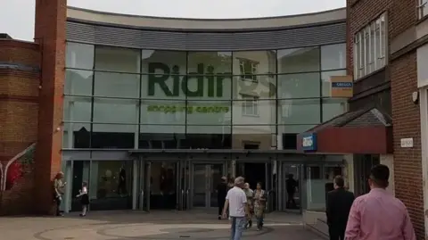LDRS The entrance of a building with glass windows and doors with people outside. There is a sign in large green writing which says 'Ridings shopping centre'.
