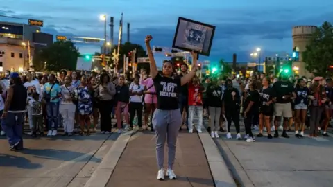 A woman in a black shirt that reads "we been not breathing" holds her fist and her hand in the air while other protesters stand in the background