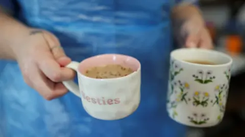 The image shows a close-up of a person holding two mugs filled with hot drinks, likely tea or coffee.