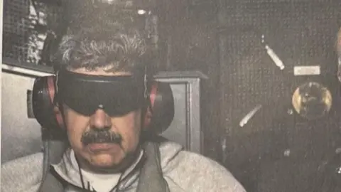 Nicolás Maduro shown after his arrest, wearing headphones and a blindfold, grey zipped jumper