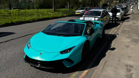 A teal-coloured Lamborghini on the side of a road with two police vehicles in the background