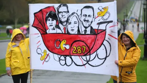PA Media Two women protesting at Stormont hold a placard depicting Arlene Foster, Sir Jeffrey Donaldson, Michelle O'Neill and Conor Murphy in a pram