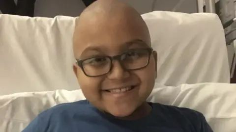 David Grist A little boy with no hair and dark rimmed glasses wears a navy t-shirt and smiles broadly at the camera from a hospital bed