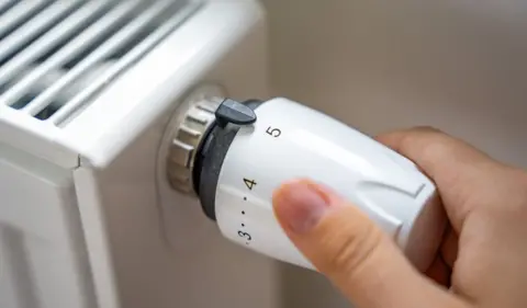 A close-up image of a person's hand turning a white radiator thermostat valve with numbers to the highest setting.