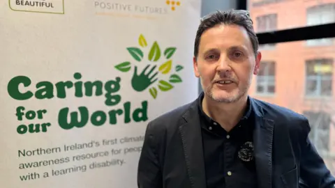 Scott Howes is wearing a black shirt with a black blazer on top. He has short dark hair and a grey beard. He is pictured beside a banner that reads 'Caring for our World' with a green hand and leaves beside it. Behind him is a window. 