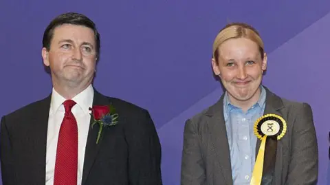 Getty Images Douglas Alexander with a sad face is wearing a dark suit, white shirt and red tie and has a red rose as a buttonhole. He is standing next to Mhair Black who has a happy face. She is wearing a grey jacket and blue shirt. she is wearing a yellow and black SNP rosette