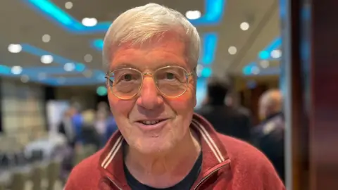 A man with short white hair and glasses is wearing a red zip-up sweater over a black shirt. The background shows an indoor setting with blue ceiling lights and several people in the distance.