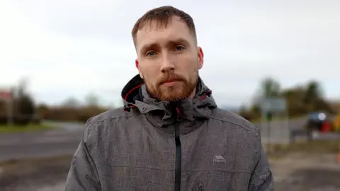 A man with short reddish hair and a beard is standing outside in front of a road. He is wearing a grey hooded jacket. The road and countryside behind him is out of focus. 