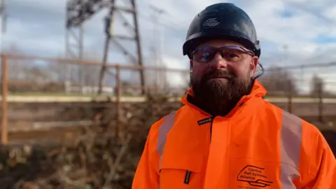 Paul wears a black hardhat, safety glasses and an orange hi-vis jacket