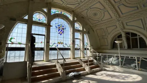 A semi-circular mezzanine floor window with a stained glass crest in the middle looks out on to Torquay harbour. The stained glass windows frame has corroded but light still pours through it casting shadows of a crest on the floor.