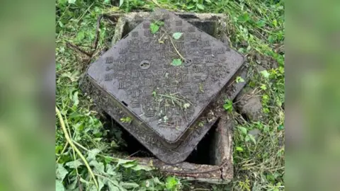 LDRS The manhole and its metal cover, which has been placed across the manhole rather than secured on. Grass surrounds the manhole.