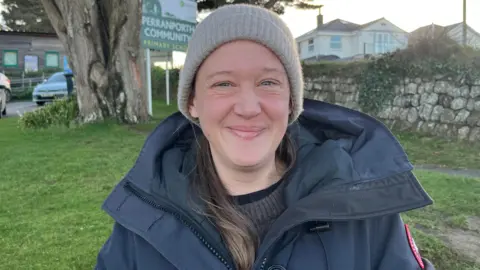 Sophie O'Shea wearing a hat and coat and smiling. She is stood near a tree and a sign which says Perranporth Community.