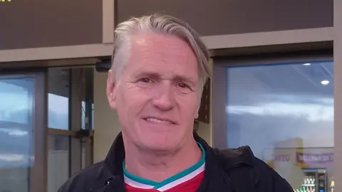 Mr. Boyd has a short gray hair and wears a Liverpool soccer club and a black coat. There is a black luggage strap on your shoulder. He is smiling at the camera.