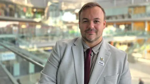 Terry Jermy is wearing a light grey suit jacket over a grey shirt, with a purple tie.