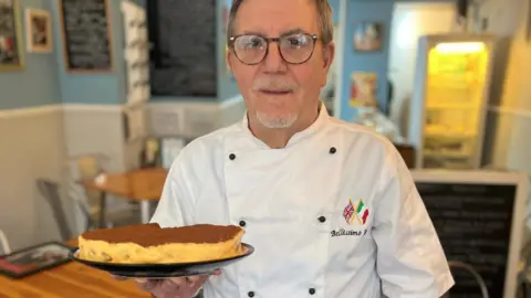 BBC Giovanni Scidetta, known to regulars as John, dressed in chef's whites and holding a plate of tiramisu
