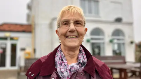 Joan Pickering stands outside Lynwood Miners Convalescent Home in Scarborough. She wears a burgundy, corduroy coat and a pink and white patterned scarf. She has cropped blonde hair and wear glasses.