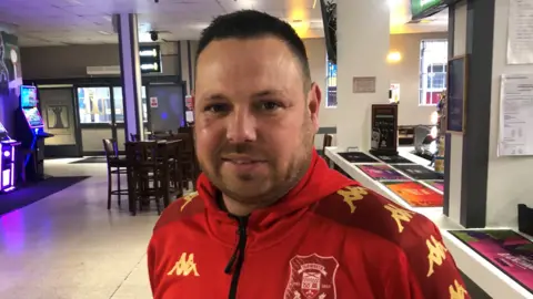 Ben Cooke is wearing a red football top, whilst stood at the bar. In the background is a slot machine and seating.