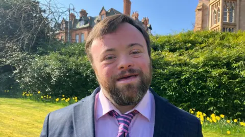 BBC James Martin smiling at the camera. He has shirt brown hair, wearing a dark blue blazer, pink shirt and pink and navy tie. A hedge, grass and daffodils are in the background.