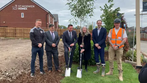 Five men and a woman stand in a line, with one of the men and the woman (both in the centre) holding spades. The man on the right is wearing a hard hat and orange hi-vis.