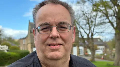 Middle-aged man with short, dark grey hair and glasses. He is standing outside. Houses and trees behind him are blurred.