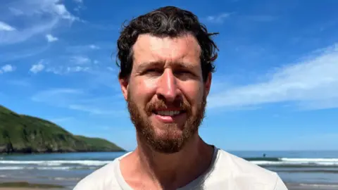 A man called Tom Franklin stands on a beach. He is smiling with his teeth showing. He has short brown hair and a beard. The sea is behind him.