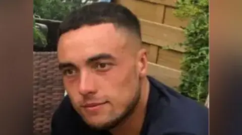 A picture of Kieran Shepherd, a who has short, black hair and stubble and is wearing a blue top. It is a close up of his face. There is a fence behind him and part of a hedge is visible on the right hand side.