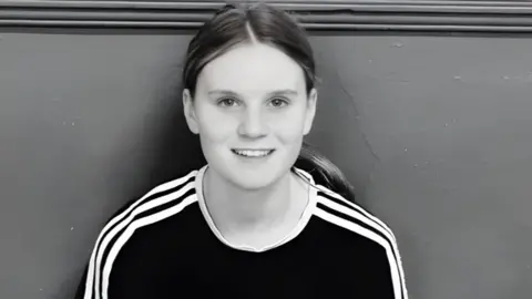 FAMILY HANDOUT Black and white close-up of Holly Newton, smiling at the camera. Her long hair is tied back in a ponytail and she is wearing a dark t-shirt with lighter stripes on the arms and shoulder.