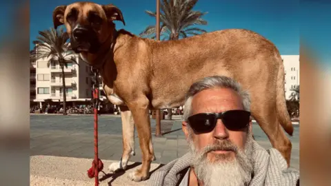 Jon Moss Jon Moss looks at the camera, he is wearing black sunglasses and has a long white beard and moustache. He has short cropped grey hair and is taking a selfie. Behind him, his Spanish Mastin dog Ripley stands side on. He has a light brown coat and is looking at the camera. He is stood on a wall. Behind the pair is a blue sky and palm trees and white buildings. 