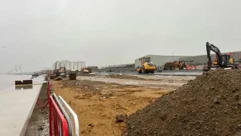 Andrew Turner/BBC Concrete river wall capping on the left, moving through uneven sand and soil with level ground and earth moving equipment in the background