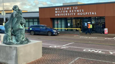 Amy Holmes/BBC A bronze sculpture of three abstract figures stands near the entrance of Milton Keynes University Hospital, with cars parked nearby and a few people walking toward the building.