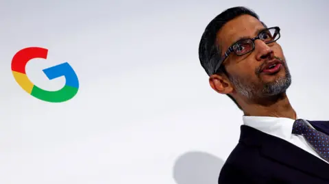Google's rainbow colored 'G' logo on a white wall behind CEO Sundar Pichai, wearing a black suit, white shirt and purple neck tie. 
