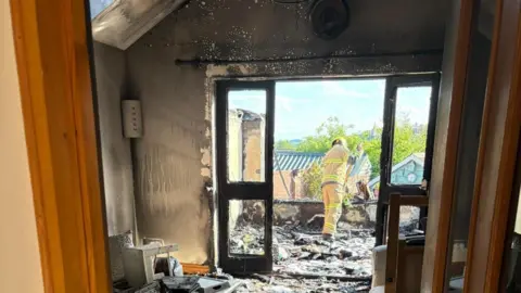 A living room that is black with fire and there are broken and burnt items scattered across the floor. Through some double doors is an area where a conservatory should be. There are no windows and there is glass and blackened debris on the floor, with a firefighter looking at it