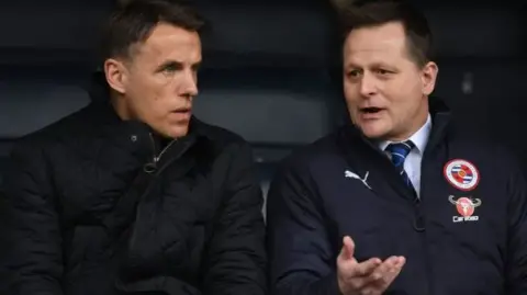 Two men sat down talking. The man on the left has short dark hair and a black jacket. The man on the right has a navy jacket, over the top of a white shirt and blue striped tie. He is gesturing to the man on the left in conversation.