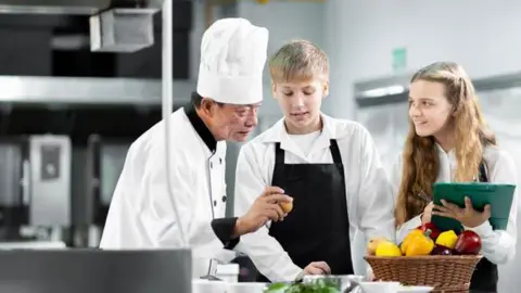 A chef, dressed in chef's whites, in a kitchen standing next to two students, also dressed in white. One of the students is a boy, the other is a girl - they both have blonde hair and are wearing black aprons. There is a basket containing fruit and vegetables in front of them.