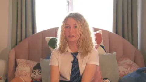 A teenage girl with long blonde curly hair sits on her bed in her school uniform. She is surrounded by pink cushions.