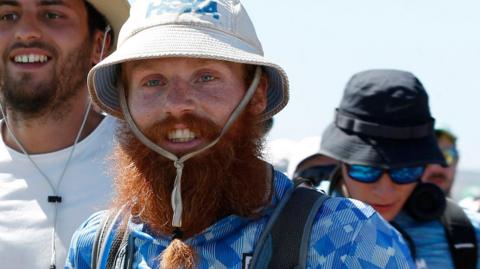 Russell Cook pictured in close up wearing a bucket hat and a blue and white England football shirt with his long red beard tied in a bunch 