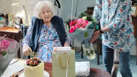 David Baker Anne Baker sits in a wheelchair and is wearing a blue dress and pearls. She isbeing presented with bouquets of flowers, a cake and gifts