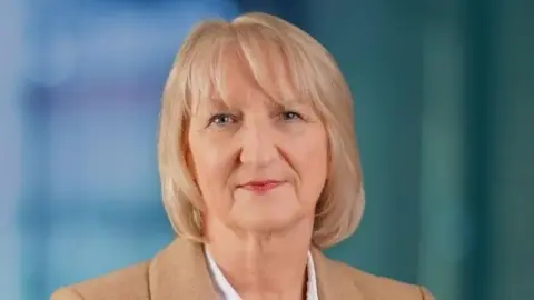 A studio-style head-and-shoulders portrait of a woman in a beige blazer and white blouse against a softly blurred background.