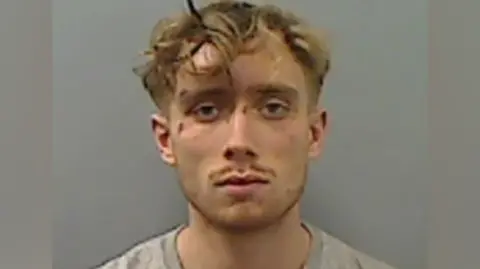 Mugshot of Callum Howe. He has blonde curly short hair and cuts to his face. He has a small moustache and slight beard and is wearing a grey t-shirt.