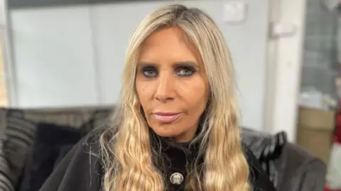 Luanne Miles is pictured with long blonde hair looks directly at the camera inside a home, with furniture and household items blurred in the background.