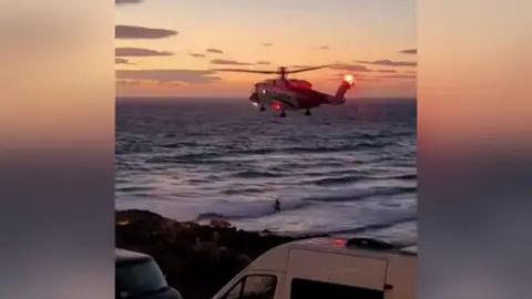 KWAN AISURU The image shows a coastguard rescue helicopter hovering low over the sea close to a shoreline at dusk. In the foreground there are two parked vans beside a coastal road or car park, while waves are breaking near the shore below and a small figure can be seen in the water or surf; the helicopter's red lights are illuminated against a sunset sky, suggesting an ongoing maritime rescue or emergency response operation near a beach.