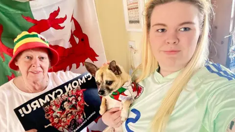 Hannah Jones A selfie of Alyson Jane and Hannah Jones and Chester, the dog. Alyson is holding a picture of the team with the words 'C'mon Cymru'. They are standing in front of the Welsh flag.